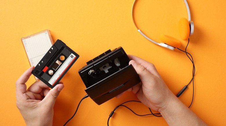 A hand holding a cassette tape with an open cassette player on an orange background.